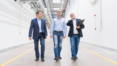 Drei Herren laufen nebeneinander durch einen Gang in einem industriellen Gebäude und halten Ausschau nach Kameras für die Videoüberwachung an der Decke.